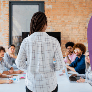 Back of a woman standing in front of colleagues sitting in a table - we can see the faces of colleagues. Main Navigating Peoples Challenges picture