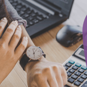 Arms of someone looking at their watch, computer and calculator on the desk.