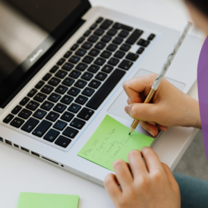 Woman's hands are on top of laptop writting a note on a post it, in order to prioritise tasks.